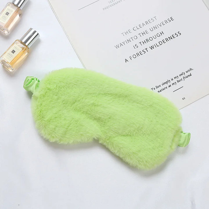 Mascherina per dormire verde in peluche su sfondo bianco, accanto a flaconi di profumo e libro. Accessorio relax, morbido e confortevole.