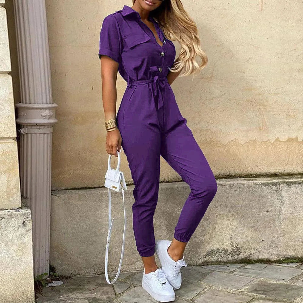 Tuta viola da donna con cintura, maniche corte e bottoni, abbinata a sneakers bianche e borsa a tracolla. Look casual ed elegante.