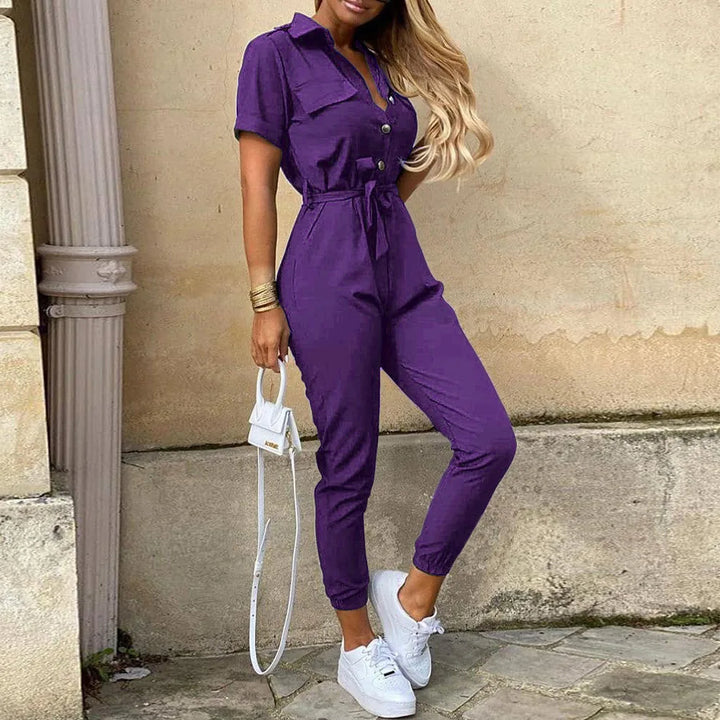 Tuta viola da donna con cintura, maniche corte e bottoni, abbinata a sneakers bianche e borsa a tracolla. Look casual ed elegante.