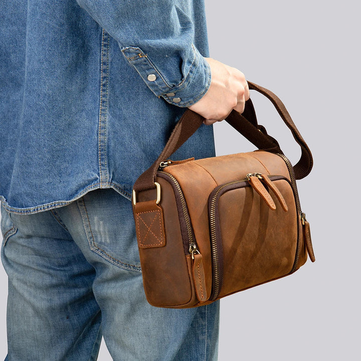 Uomo con camicia di jeans che tiene una borsa a tracolla in pelle marrone, design vintage, tasche con cerniera, moda casual, accessorio maschile.