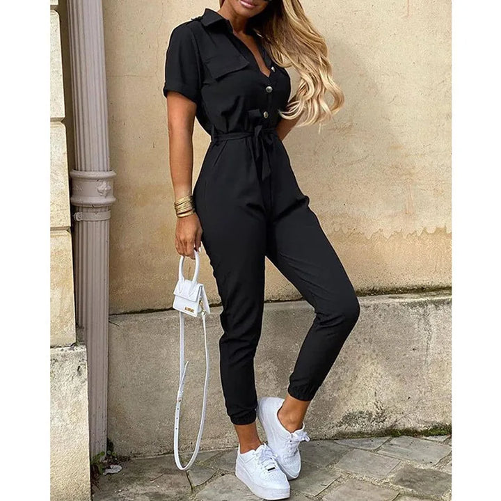 Tuta nera elegante da donna con cintura, maniche corte e bottoni, abbinata a sneakers bianche e borsa a tracolla piccola. Look casual chic.