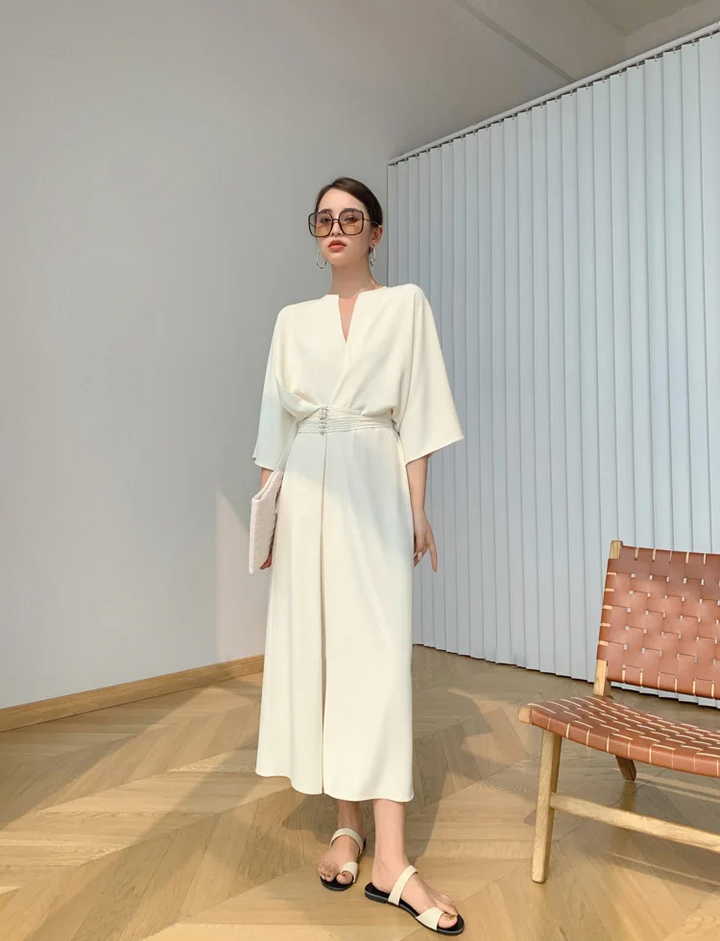 Donna in abito bianco elegante, occhiali da sole, sandali, in piedi accanto a sedia moderna. Moda estiva, stile minimalista, interni luminosi.