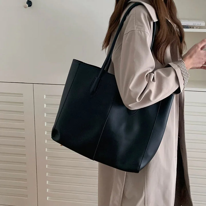 Borsa tote nera in pelle, indossata da una donna con cappotto beige. Accessorio moda elegante, ideale per shopping e uso quotidiano.