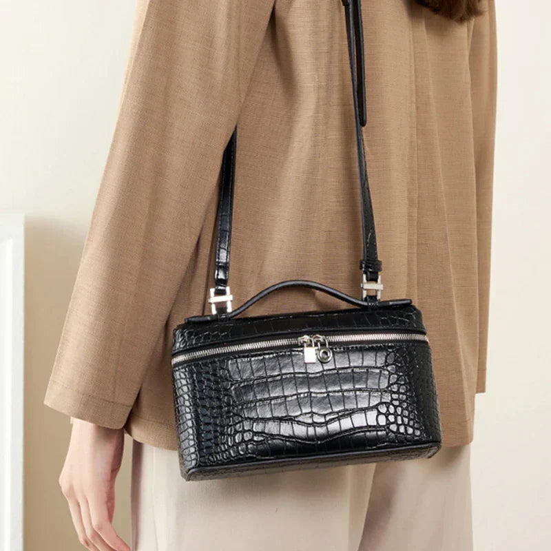Borsa a tracolla nera in pelle effetto coccodrillo, chiusura con zip e fibbia argentata, indossata su cappotto beige. Accessorio moda elegante.