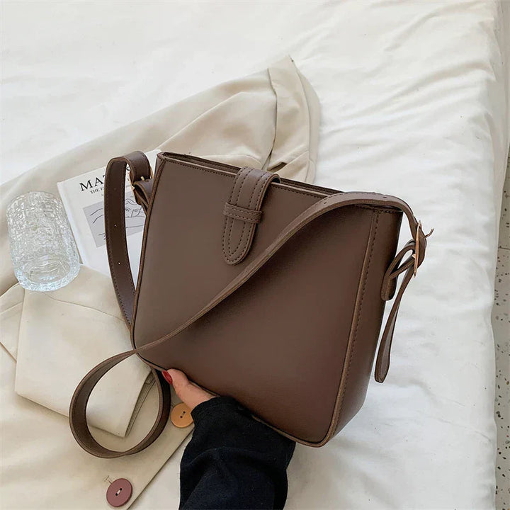Borsa a tracolla marrone in pelle su letto bianco, con rivista e bicchiere d'acqua. Elegante accessorio moda donna, design minimalista.