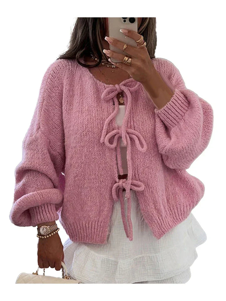 Donna con cardigan rosa in maglia morbida, maniche lunghe, fiocchi decorativi, sopra abito bianco a balze. Moda autunnale elegante e casual.