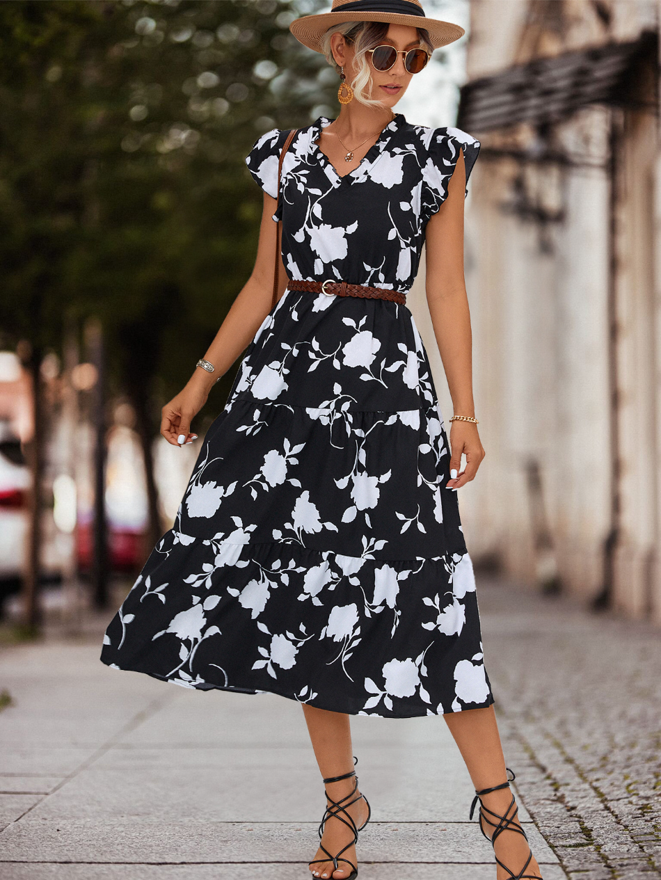 Donna con abito lungo floreale nero e bianco, cintura marrone, cappello beige e occhiali da sole. Moda estiva elegante, look casual chic.