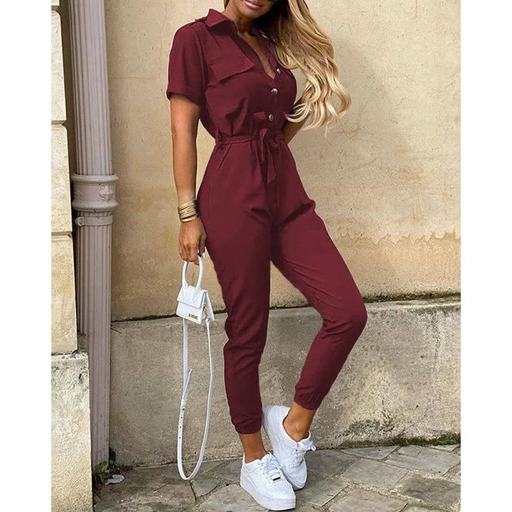 Tuta jumpsuit donna bordeaux, maniche corte, con cintura in vita, abbinata a sneakers bianche e borsa a tracolla bianca. Moda casual elegante.