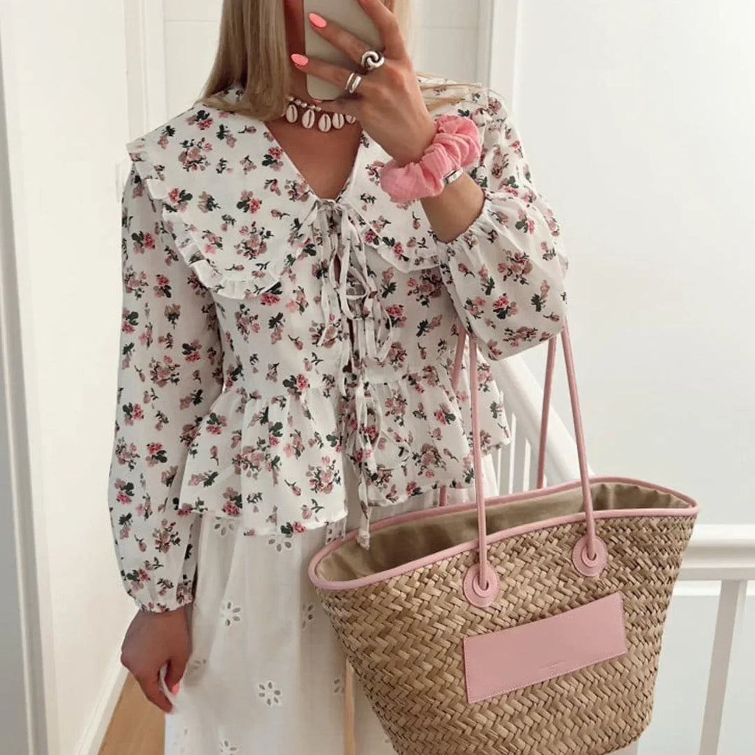Donna con camicetta floreale bianca, gonna bianca traforata, borsa di paglia con manici rosa, bracciale rosa, collana di perle, unghie rosa.