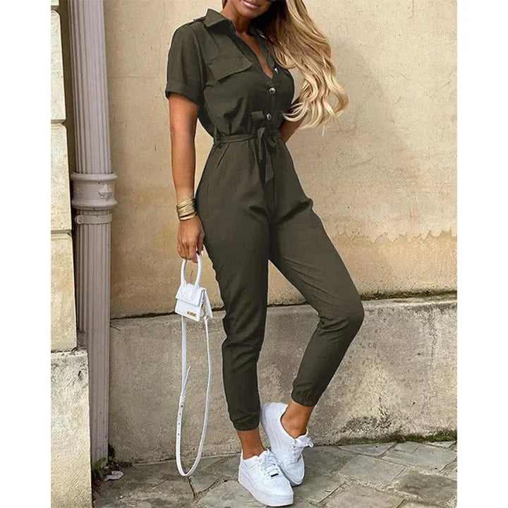 Tuta intera verde oliva da donna, maniche corte, con cintura in vita, abbinata a sneakers bianche e borsa a tracolla bianca. Moda casual elegante.