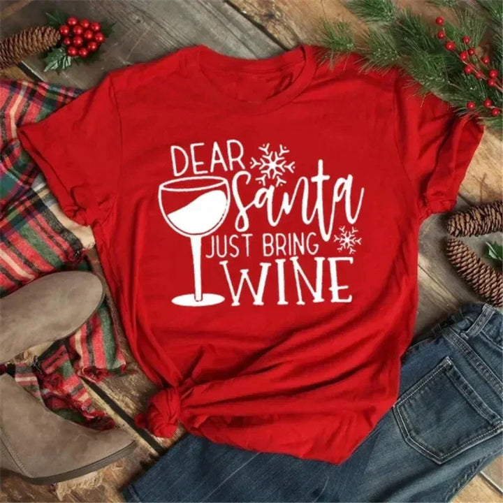 Maglietta rossa natalizia con scritta "Dear Santa Just Bring Wine", decorata con bicchiere di vino e fiocchi di neve, su sfondo di legno e decorazioni festive.