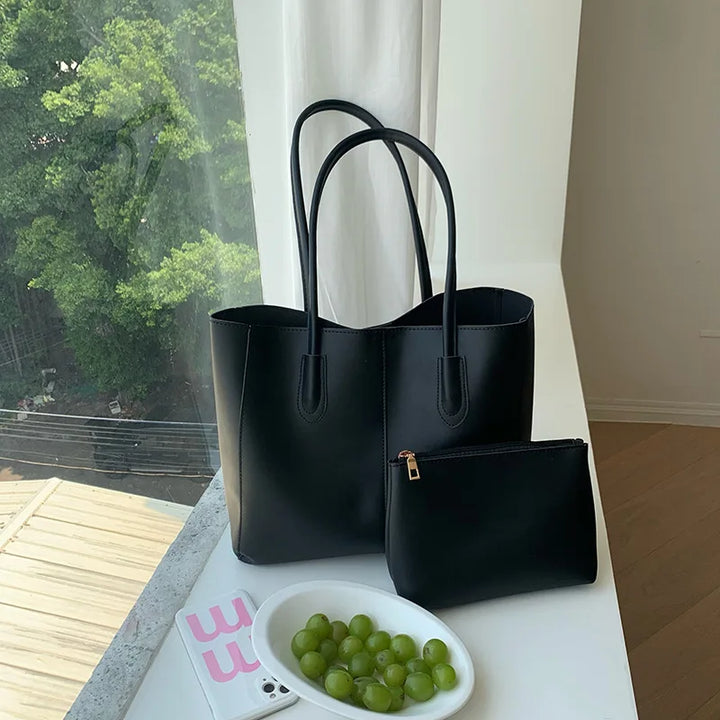 Borsa tote nera in pelle con pochette abbinata su tavolo bianco, accanto a piatto di uva verde. Design elegante, accessorio moda donna.