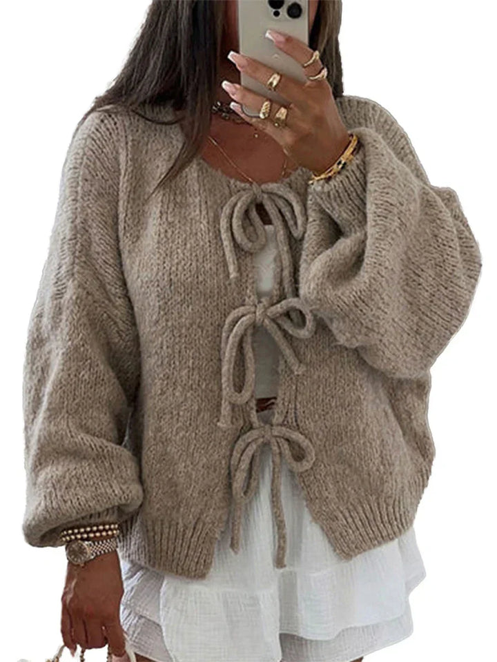 Donna con cardigan beige in maglia con fiocchi, maniche lunghe e ampie, sopra abito bianco. Moda autunnale, abbigliamento casual elegante.