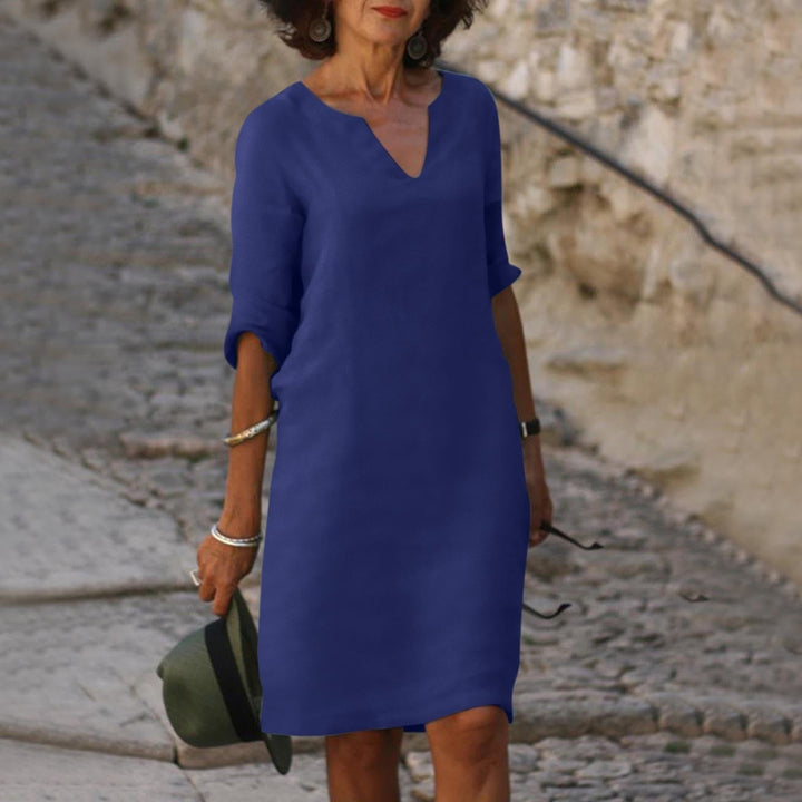 Donna con abito blu casual, maniche a tre quarti, su strada acciottolata. Moda estiva elegante, accessori argento, cappello verde in mano.