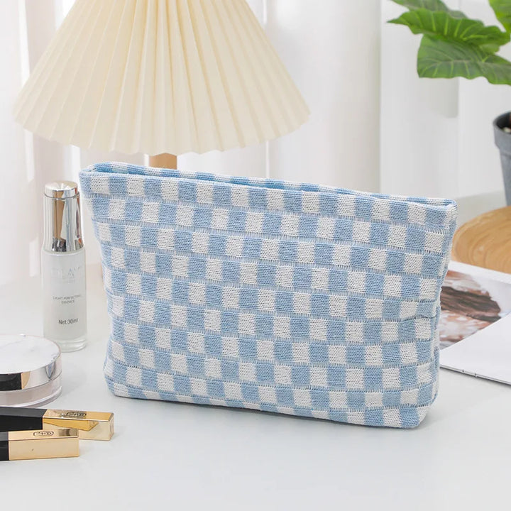 Pochette a scacchi blu e bianchi su tavolo bianco, accanto a lampada, piante e cosmetici. Accessorio moda elegante e pratico.