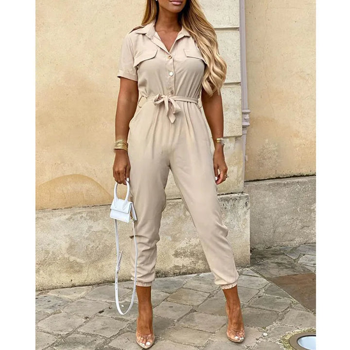 Tuta jumpsuit beige da donna, maniche corte, cintura in vita, abbinata a sandali trasparenti e borsa bianca. Look estivo elegante e casual.