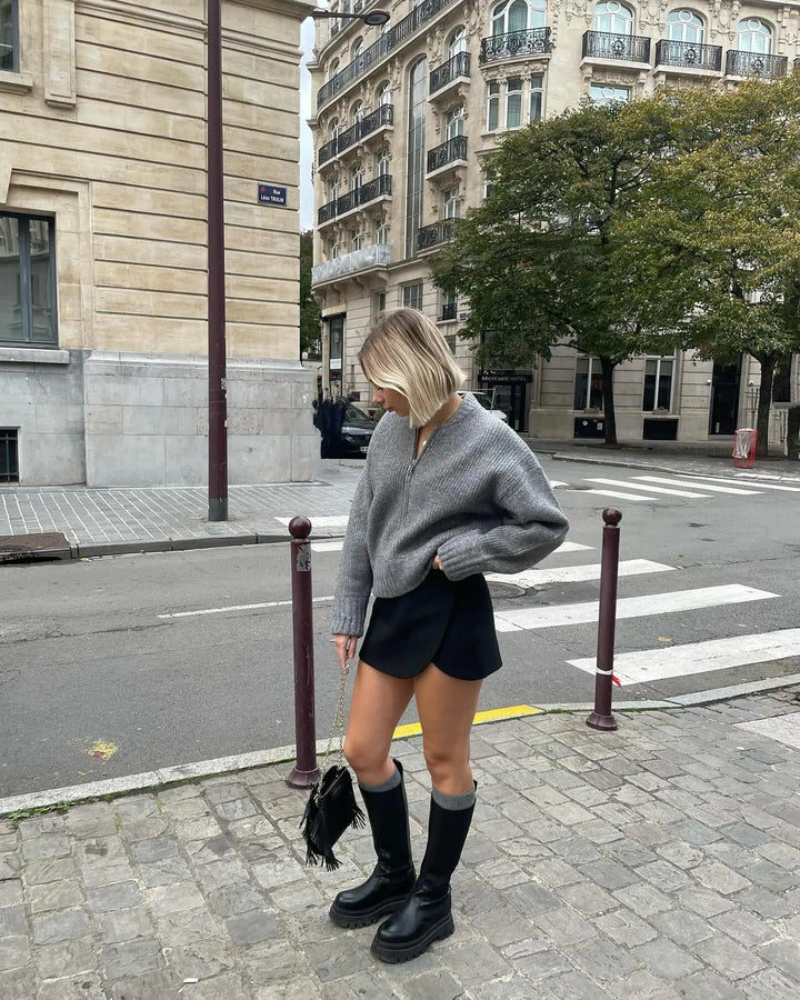 Donna con maglione grigio oversize, minigonna nera e stivali alti neri in strada cittadina. Moda autunnale urbana, look casual chic.