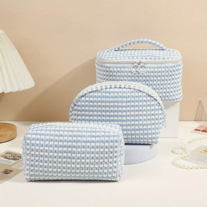 Set di tre borse da toilette in tessuto intrecciato bianco e azzurro, con cerniera e manico, su sfondo beige. Accessori da viaggio eleganti e pratici.