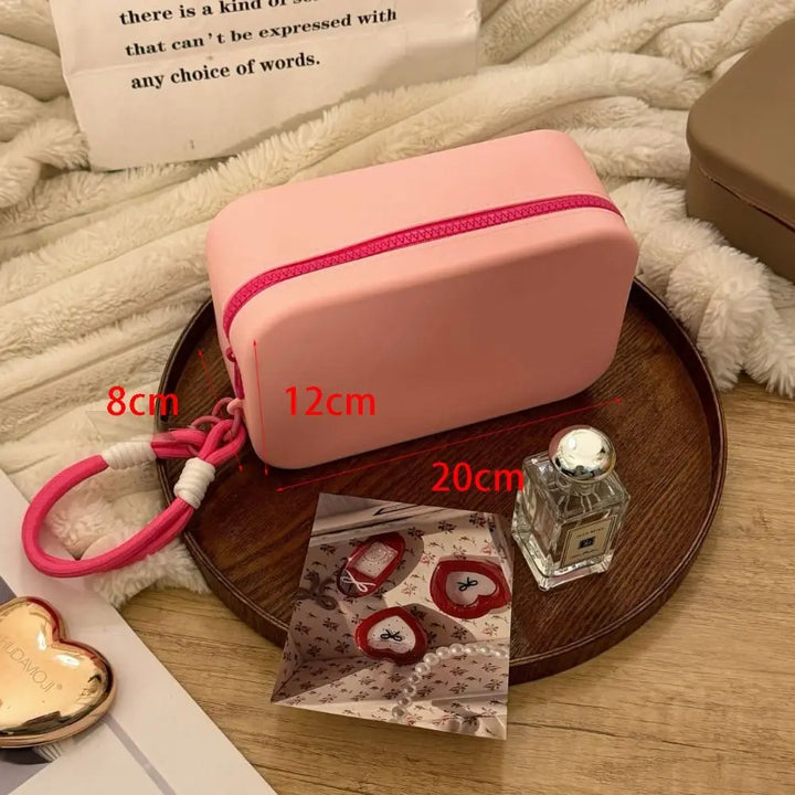 Astuccio rosa in silicone con cerniera rossa, dimensioni 20x12x8 cm, su vassoio di legno. Include profumo e accessori decorativi.