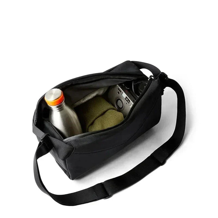 Borsa a tracolla nera aperta con bottiglia d'acqua, maglione verde e fotocamera all'interno, su sfondo bianco. Accessori viaggio, moda uomo.