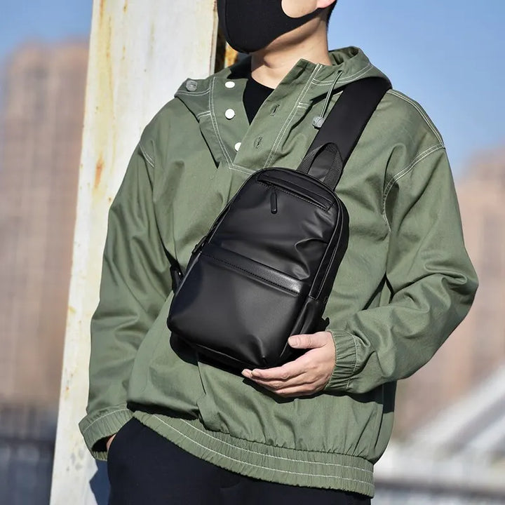 Uomo con giacca verde indossa zaino a tracolla nero in pelle, stile urbano. Accessorio moda maschile, pratico per viaggi e uso quotidiano.