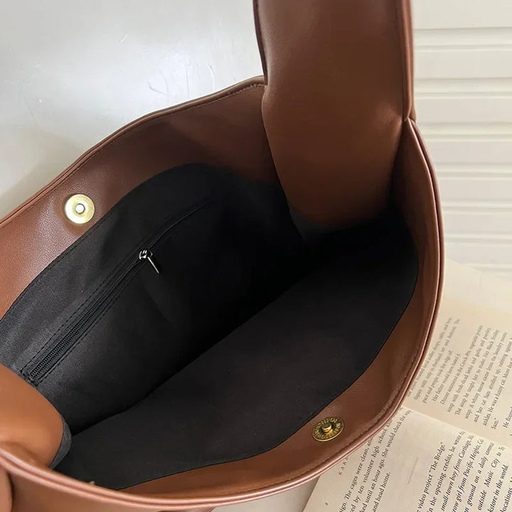 Interno di una borsa in pelle marrone con chiusura a bottone e tasca con zip, appoggiata su un libro aperto. Accessorio moda elegante e funzionale.