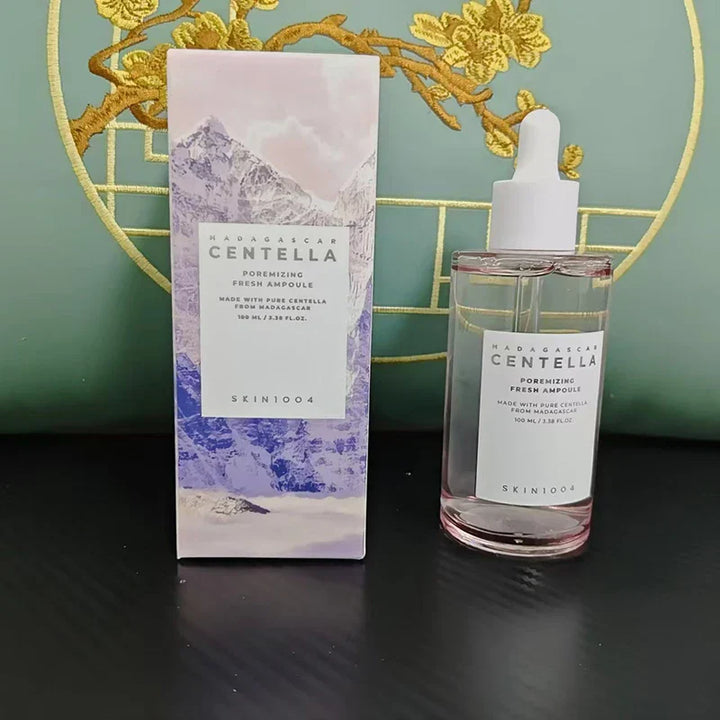 Boccetta di siero viso Skin1004 Madagascar Centella, 100 ml, con scatola decorata da montagne e fiori gialli, su sfondo verde.
