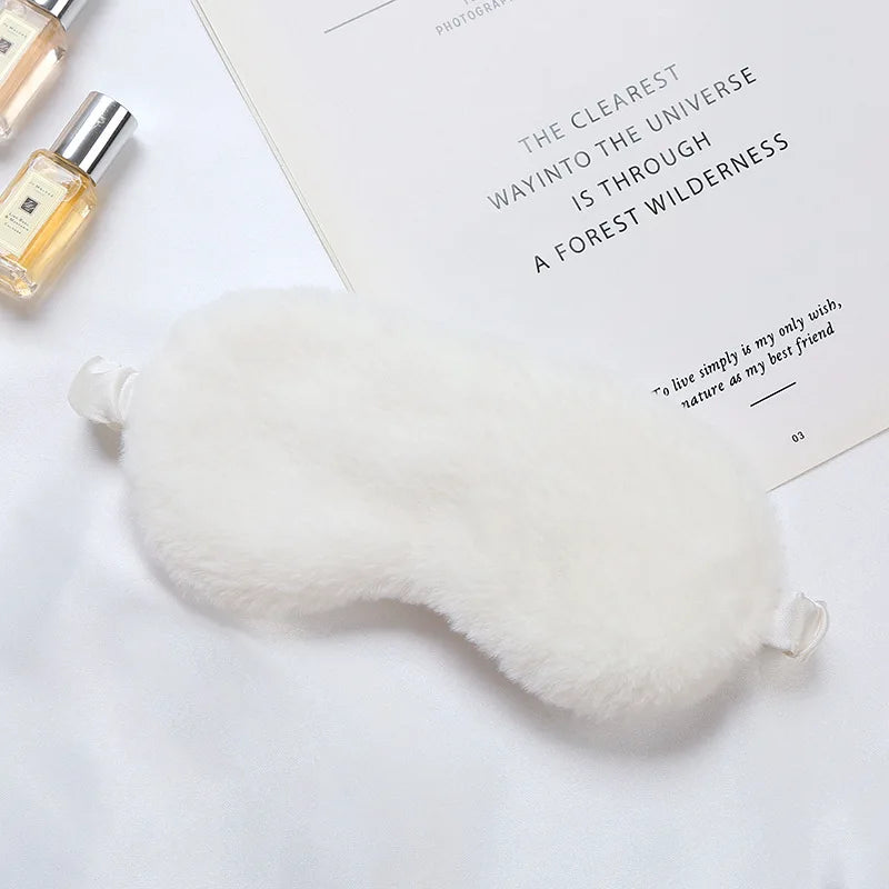 Maschera per dormire in pelliccia bianca su sfondo bianco, accanto a flaconi di profumo e un libro. Accessorio relax, comfort, sonno riposante.