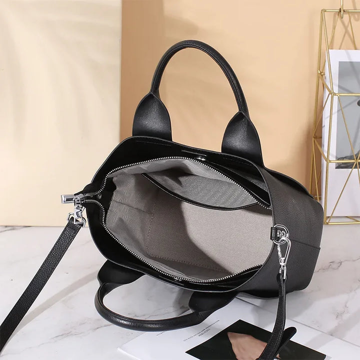 Borsa a mano nera in pelle con interno beige, cerniera aperta, manici robusti e tracolla regolabile, su sfondo chiaro. Accessorio moda elegante.