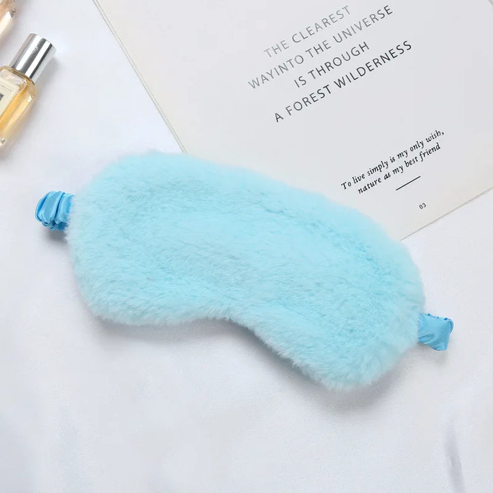 Mascherina per dormire in pelliccia sintetica azzurra su sfondo bianco, accanto a un libro e una bottiglia di profumo. Accessorio relax e benessere.