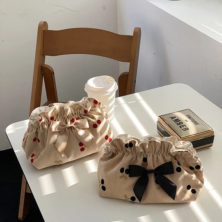 Due borse in tessuto beige con fiocchi, una con motivi a ciliegia rossa e l'altra a pois neri, su tavolo bianco accanto a sedia e bevanda.