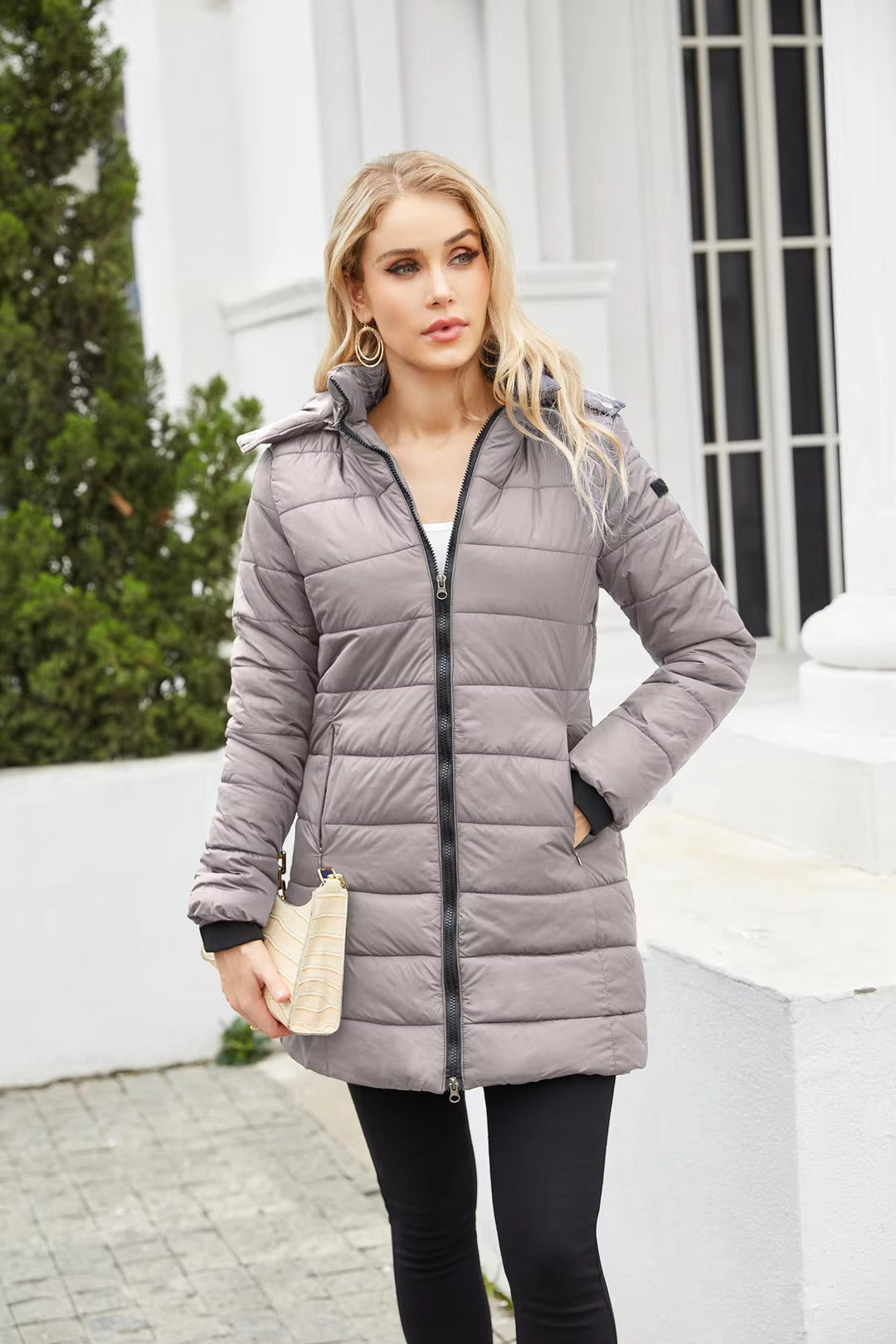 Donna con cappotto imbottito grigio, zip frontale, cappuccio, tasche laterali, posa all'aperto. Moda invernale, abbigliamento caldo, stile casual.
