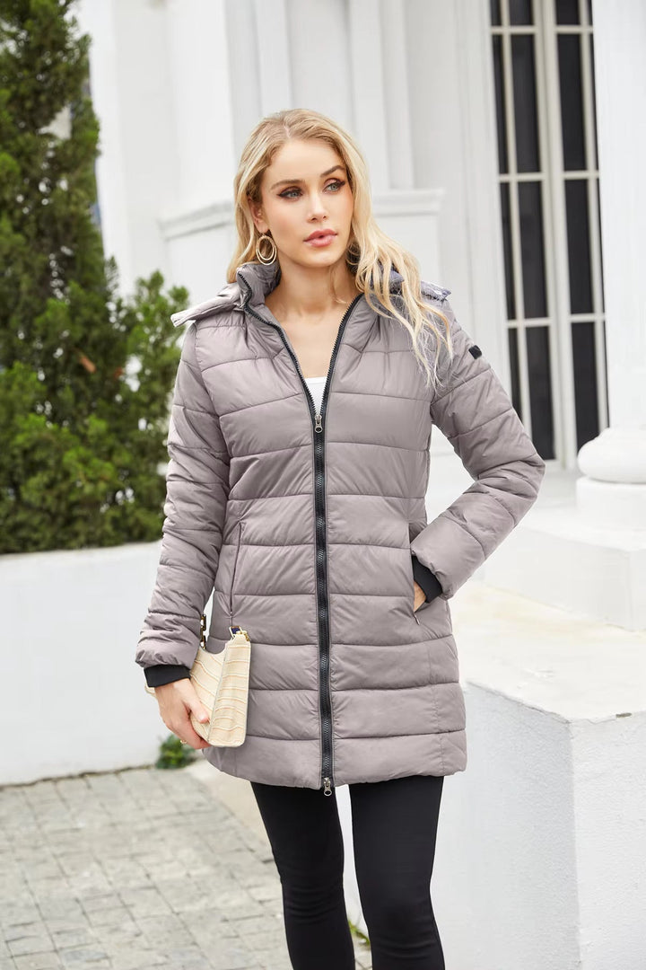 Donna con cappotto imbottito grigio, zip frontale, cappuccio, tasche laterali, posa all'aperto. Moda invernale, abbigliamento caldo, stile casual.