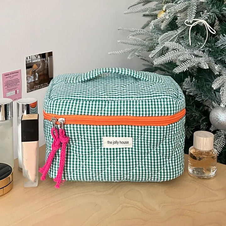 Borsa cosmetica a quadretti verdi con cerniera arancione su tavolo di legno, accanto a prodotti di bellezza e albero di Natale decorato.