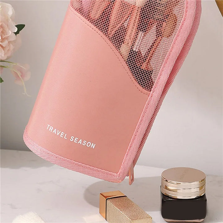Custodia rosa per pennelli trucco con rete, scritta "Travel Season", accanto a rossetto e crema viso su tavolo bianco. Accessori beauty viaggio.