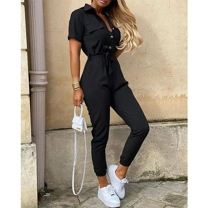 Tuta nera elegante da donna con cintura, maniche corte e bottoni, abbinata a sneakers bianche e borsa a tracolla piccola. Look casual chic.