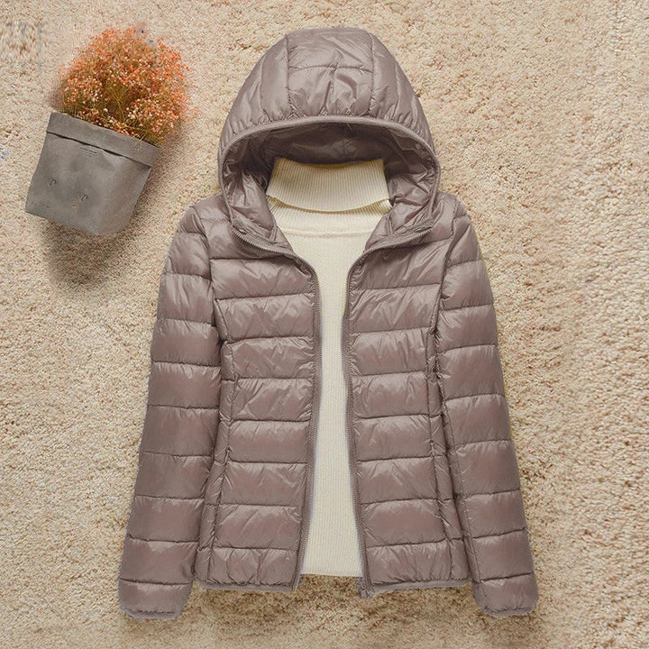 Giacca invernale imbottita da donna con cappuccio, colore marrone chiaro, su sfondo beige. Elegante e calda, ideale per la stagione fredda.