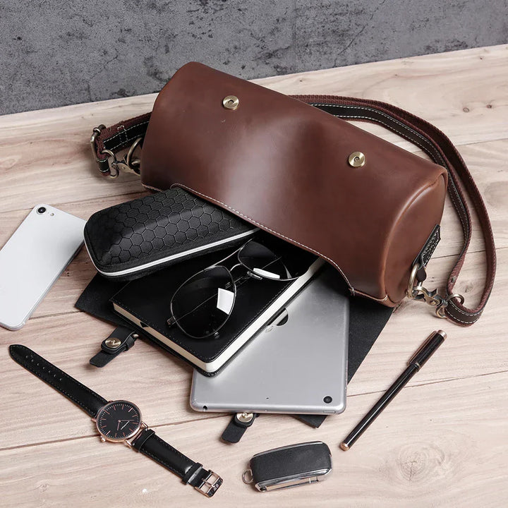 Borsa a tracolla in pelle marrone su tavolo di legno, contenente occhiali, smartphone, tablet, orologio e penna. Accessori moda eleganti.