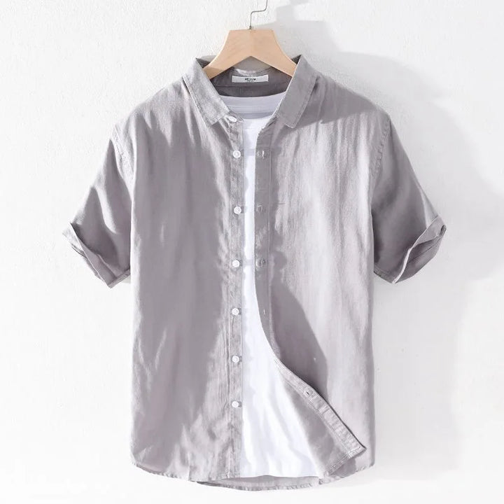 Camicia grigia a maniche corte in cotone, stile casual, con colletto classico e bottoni bianchi, appesa su gruccia di legno.