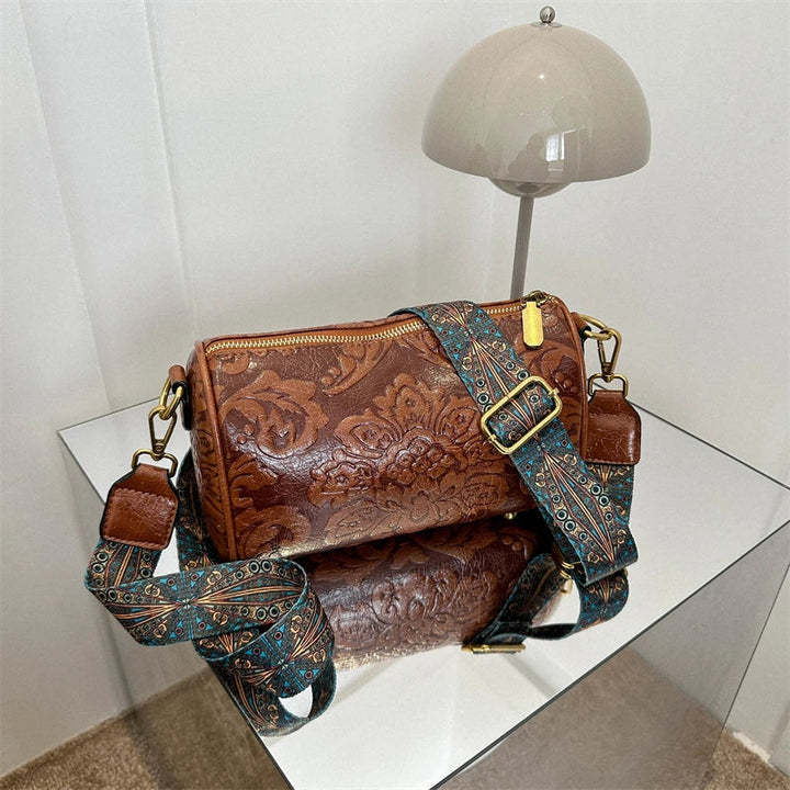 Borsa a tracolla in pelle marrone con motivo floreale, cinturino regolabile blu decorato, su tavolo a specchio, lampada moderna sullo sfondo.