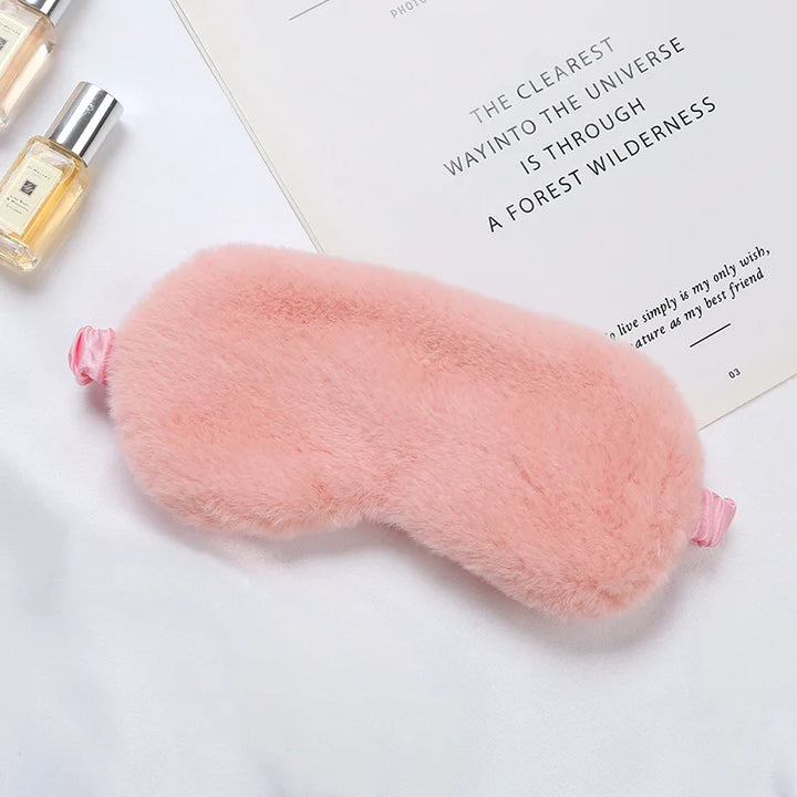 Mascherina per dormire in peluche rosa su sfondo bianco, accanto a flaconi di profumo e libro. Accessorio per il sonno, morbido e confortevole.