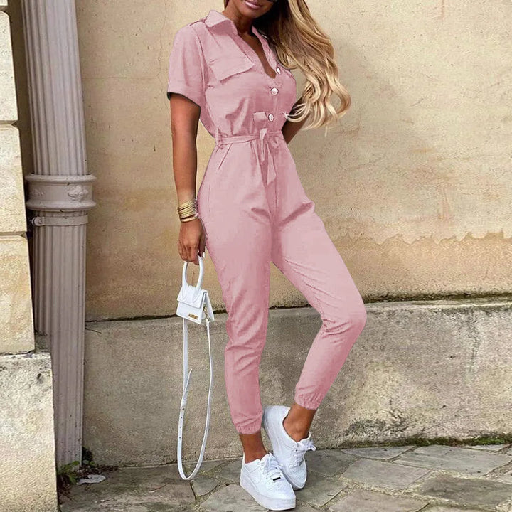 Tuta jumpsuit rosa donna, maniche corte, cintura in vita, abbinata a sneakers bianche e borsa piccola. Look casual chic estivo.