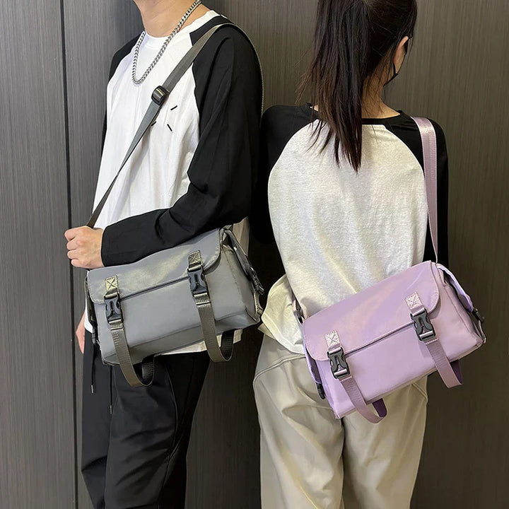 Due persone con borse a tracolla unisex, una grigia e una lilla, indossano maglie bianche e nere. Stile casual, moda urbana.