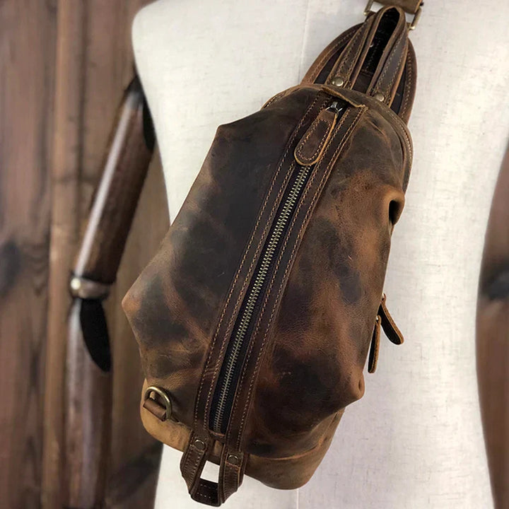 Zaino a tracolla in pelle vintage marrone, design unisex, con cerniera centrale e dettagli in metallo, ideale per viaggi e uso quotidiano.