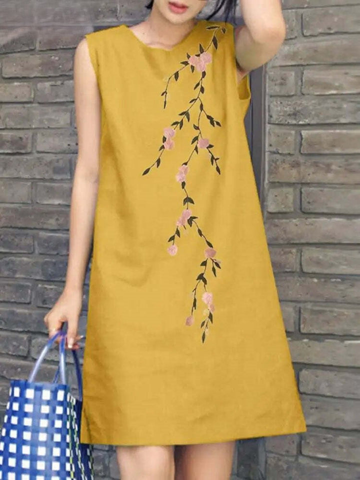 Abito estivo giallo senape senza maniche con motivo floreale ricamato, donna con borsa a quadri blu, sfondo muro di mattoni.