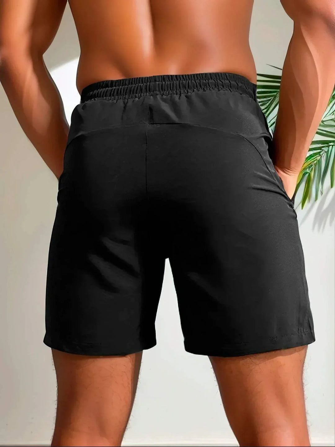 Shorts sportivi neri da uomo, tessuto leggero e traspirante, elastico in vita, ideali per allenamento e fitness. Sfondo con pianta verde.