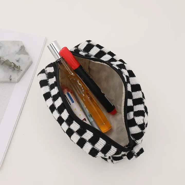 Astuccio a scacchi bianco e nero aperto con penne e spray profumato su sfondo bianco. Accessorio moda, organizer da borsa, idea regalo pratica.