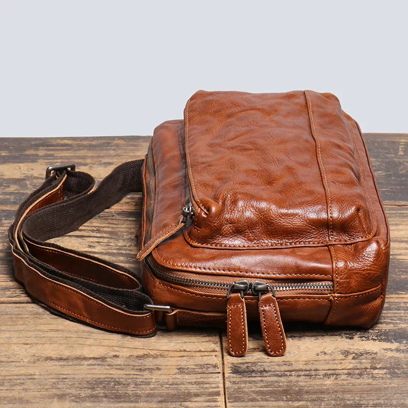 Borsa a tracolla in pelle marrone vintage su tavolo di legno, con cerniere robuste e tracolla regolabile. Accessorio moda uomo elegante e pratico.