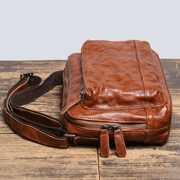 Borsa a tracolla in pelle marrone vintage su tavolo di legno, con cerniere robuste e tracolla regolabile. Accessorio moda uomo elegante e pratico.