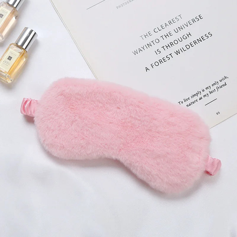 Mascherina per dormire in peluche rosa su sfondo bianco con flaconi di profumo e libro. Accessorio per il sonno, morbido e confortevole.